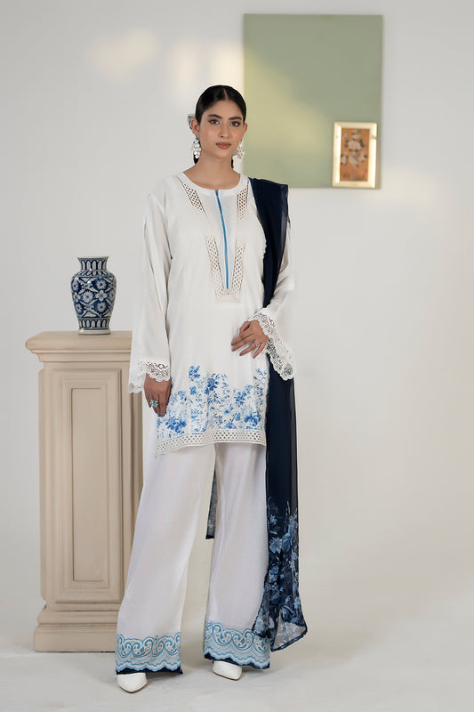 Woman wearing a white and blue embroidered outfit in a minimalistic setting.