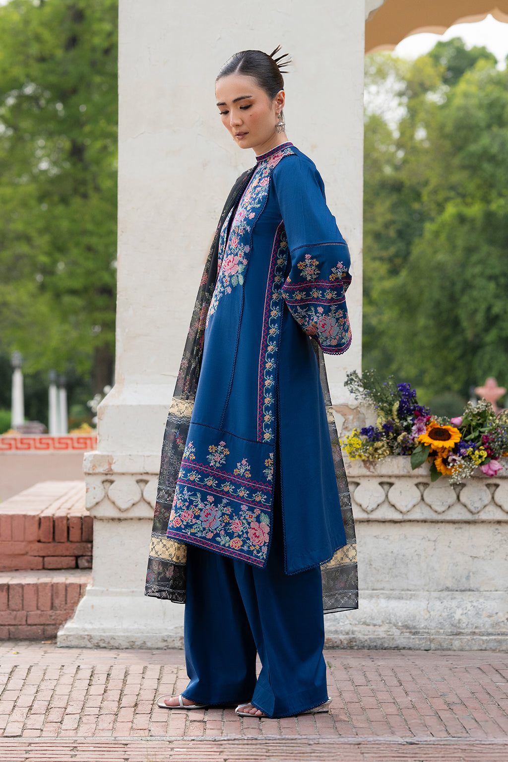 Woman in traditional blue outfit with floral embroidery standing in a garden.