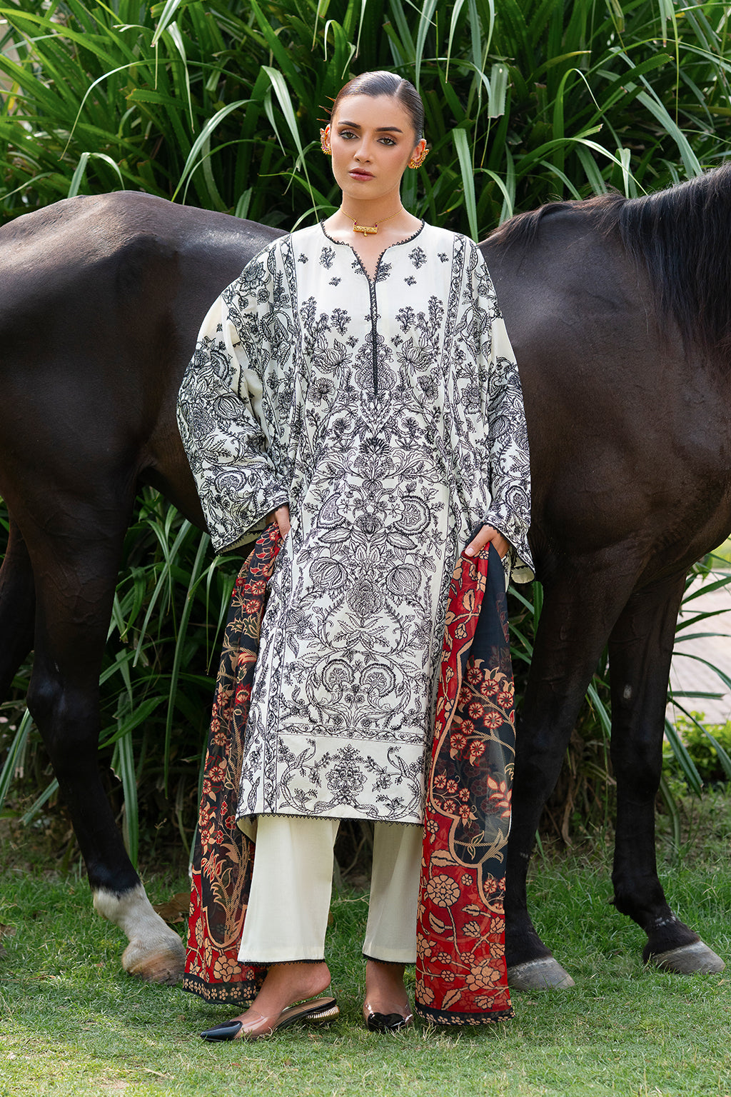 A woman standing in front of a horse, wearing a white and black embroidered ensemble with a multicolored chiffon dupatta.