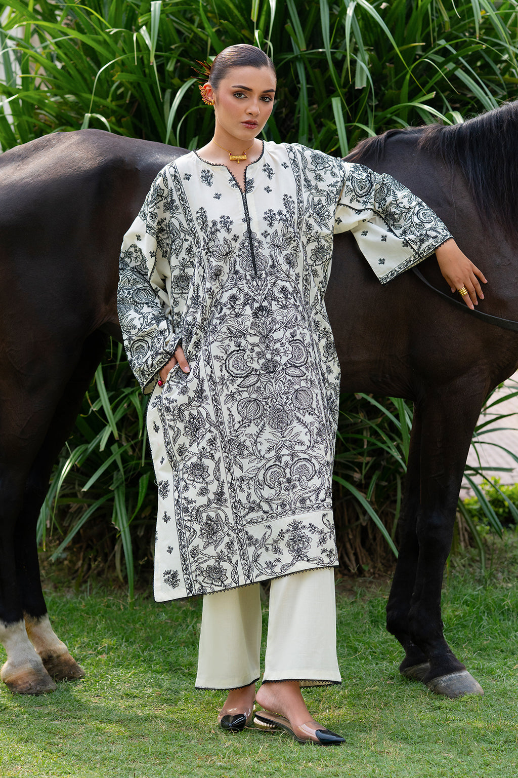 Woman in patterned kurta and white pants standing with a horse in a grassy area.
