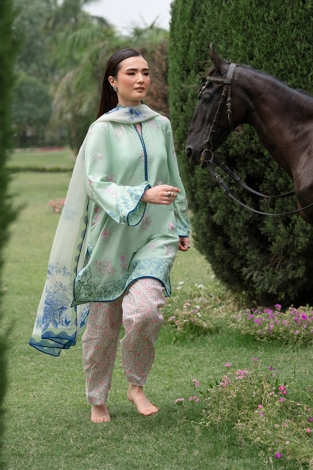 Woman in light green outfit walking with a horse in a garden.