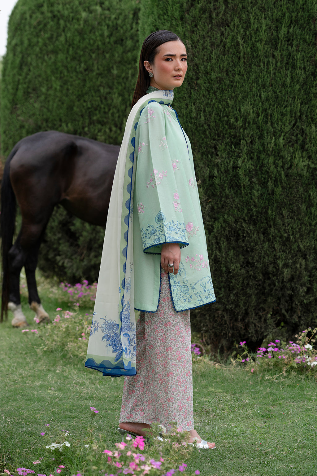 Woman in light green embroidered coat standing outdoors with a horse in the background.