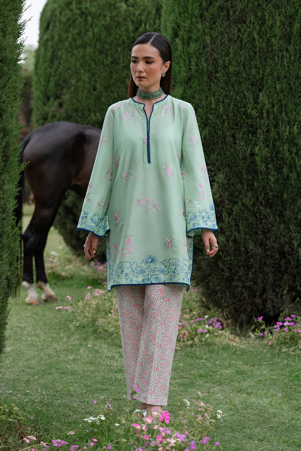 Woman in light green dress walking in a garden with a horse in the background.