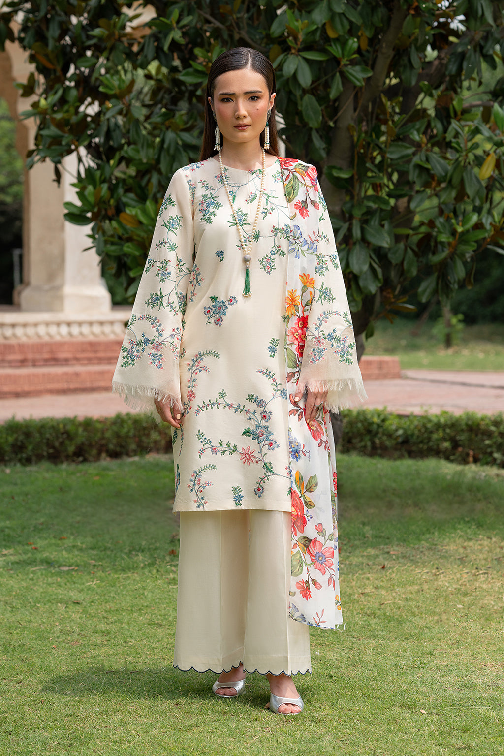 A woman wearing an off-white ensemble with floral embroidery on the front and sleeves, paired with a chiffon dupatta and dyed trousers.