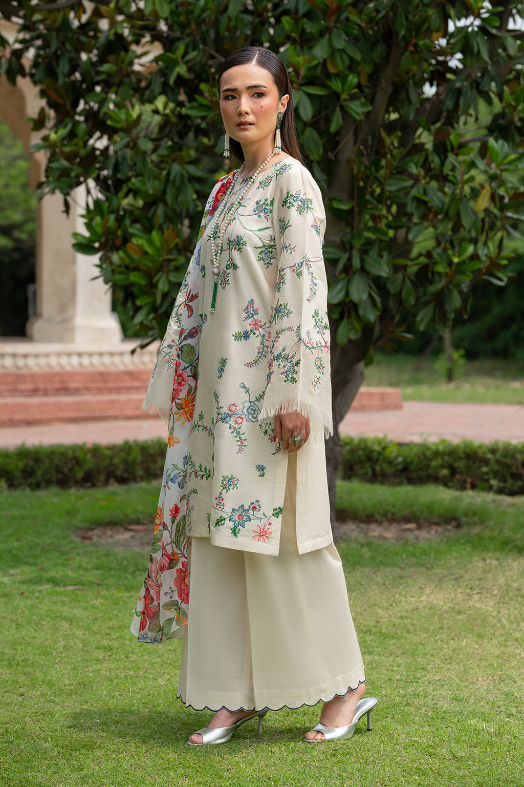 Woman in floral dress and beige pants standing on grass with trees in the background.