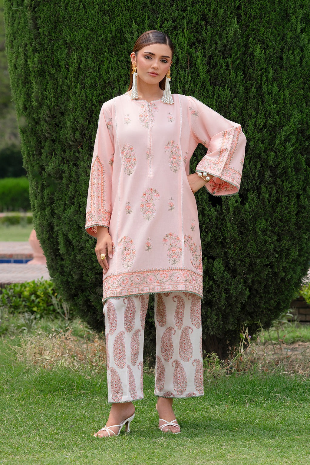Woman in a pink traditional outfit standing outdoors with greenery in the background.