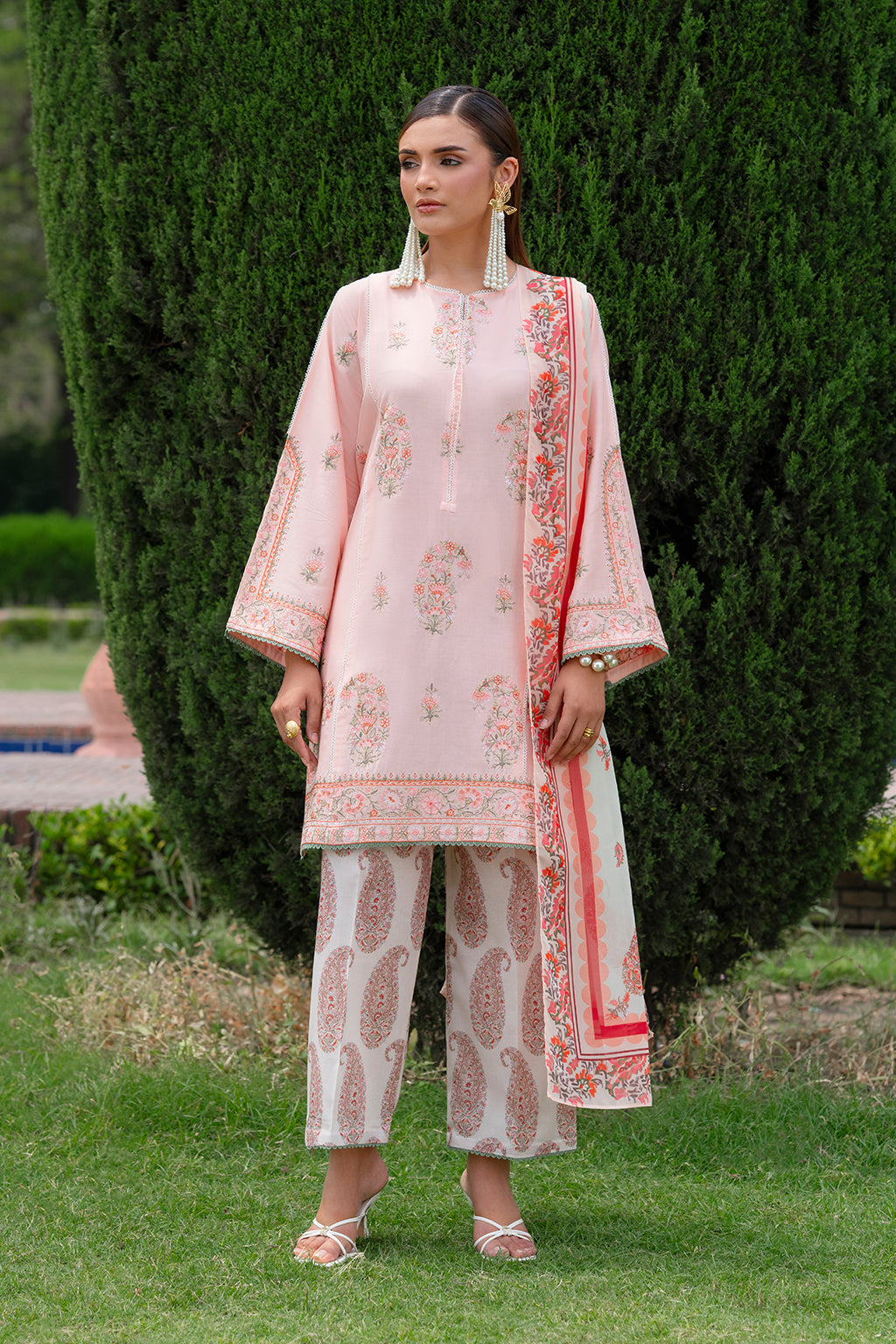 A woman wearing a baby pink ensemble with intricate embroidery, a digital printed silk dupatta, and matching printed lawn trousers, standing in a garden setting.
