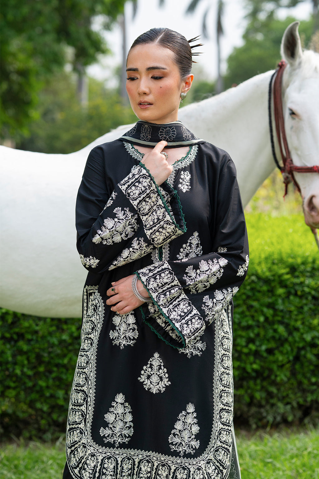 Woman in black and silver embroidered outfit standing with a horse in the background.