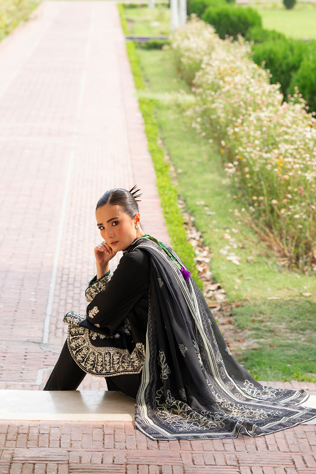 Woman in black and silver traditional outfit sitting on a path in a garden.