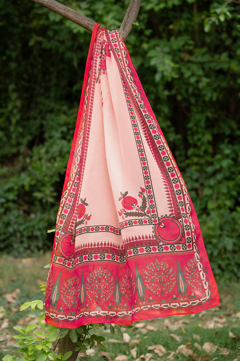 Traditional red and beige patterned fabric hanging on a tree branch with greenery in the background.